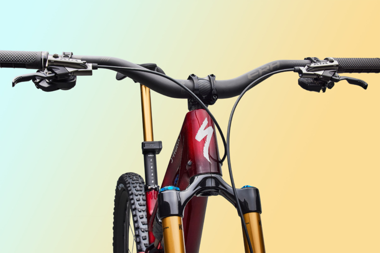 Close-up view of a modern mountain bike's handlebars, featuring textured grips, gear shifters, and hydraulic brake levers, with a sleek red and black bike frame in the background. The image has a soft gradient background in shades of blue and yellow.