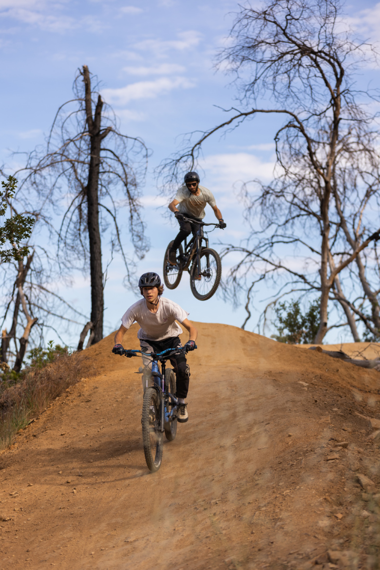 Two mountain bikers are riding on a dirt trail, one jumping off a ramp while the other rides close to the ground. The scene features dry vegetation and charred trees in the background, under a partly cloudy blue sky.