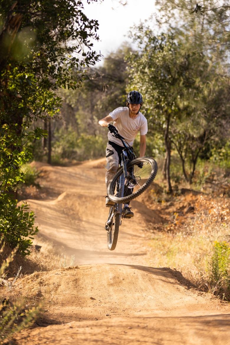 A person riding a mountain bike performs a jump on a dirt trail surrounded by greenery. The rider is wearing a helmet and casual clothing, with the bike's front wheel lifted off the ground as they navigate the jump. Dust is kicking up around the bike, indicating a dynamic movement. The background features a natural landscape with trees and shrubs.