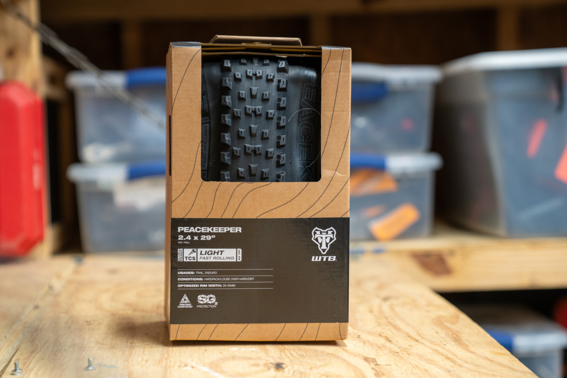 A boxed bicycle tire labeled 'Peacekeeper 2.4 x 29 inches' with descriptions of its features, including 'Light' and 'Fast Rolling.' The tire is partially visible through a clear window in the box, which is placed on a wooden surface with storage bins in the background.
