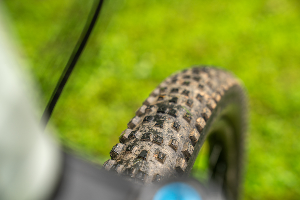 Close-up view of a mountain bike tire with a heavily textured tread, showing dirt and mud, set against a blurred green background.