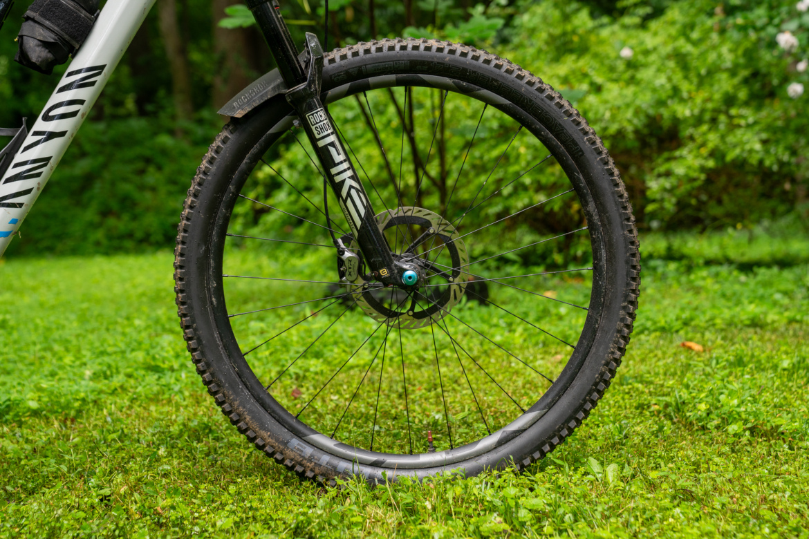 Close-up of a mountain bike front wheel on a grassy surface, showcasing a black tire with a textured tread pattern, a wheel hub with a blue accent, and a suspension fork labeled "Pike." The background features lush green foliage.
