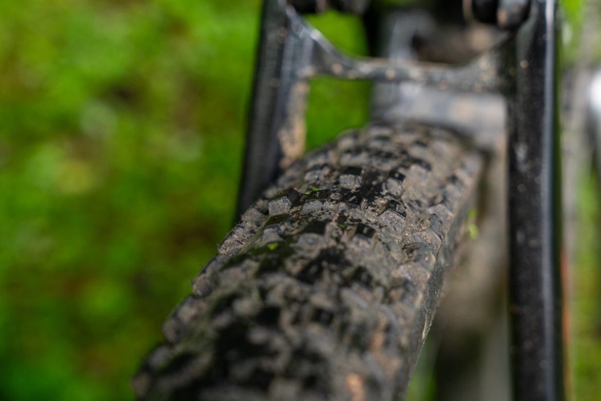 Close-up of a mountain bike tire with visible tread patterns, showing dirt and mud, against a blurred green background, likely indicating a recent ride on a trail or rough terrain.