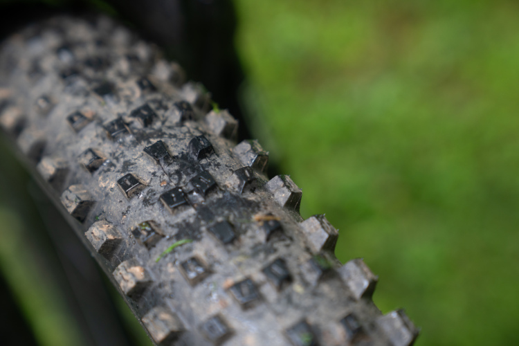 Close-up of a WTB Peacemaker mountain bike tire, showcasing knobby tread patterns and dirt accumulated on the surface, set against a blurred green background.