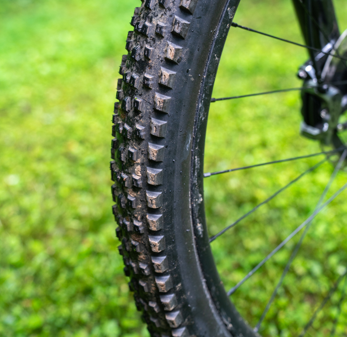 Close-up of a mountain bike tire with a rugged tread, showing visible dirt and moisture, set against a grassy background.