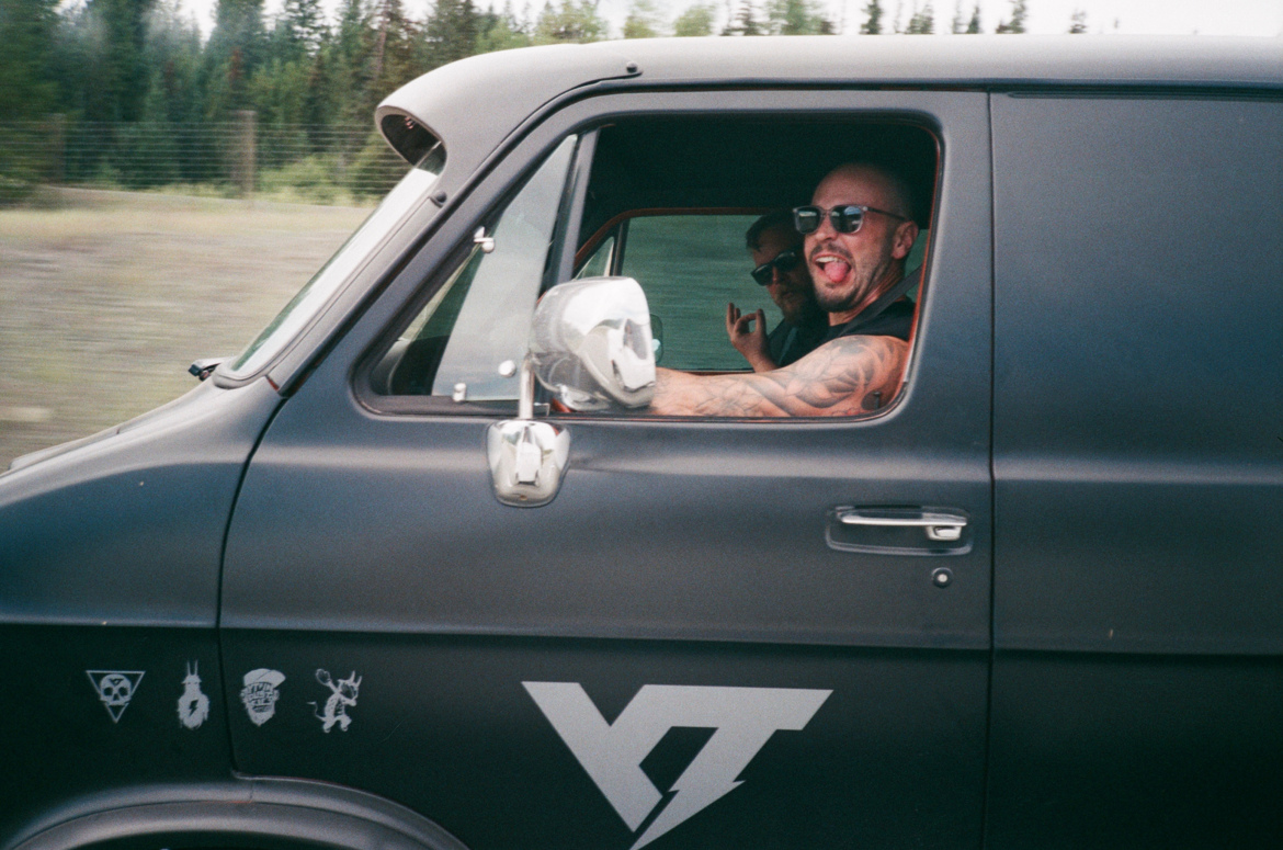 Two men are seen inside a black van with various decals on the side. The driver is wearing sunglasses and sticking out his tongue, while the passenger, also in sunglasses, is making a hand gesture. The background includes greenery and trees, suggesting they are driving through a natural setting.