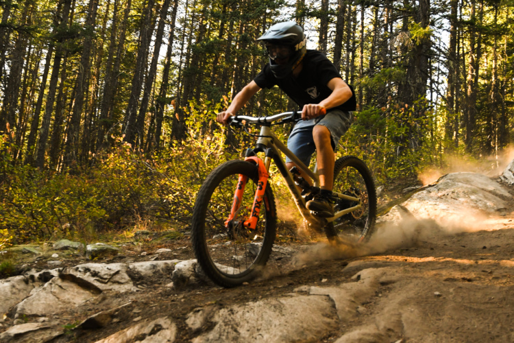 A mountain biker navigating a rocky trail in a forested area, kicking up dust as they ride. The rider is wearing a helmet and is dressed casually in a t-shirt and shorts. Sunlight filters through the trees, illuminating the scene.