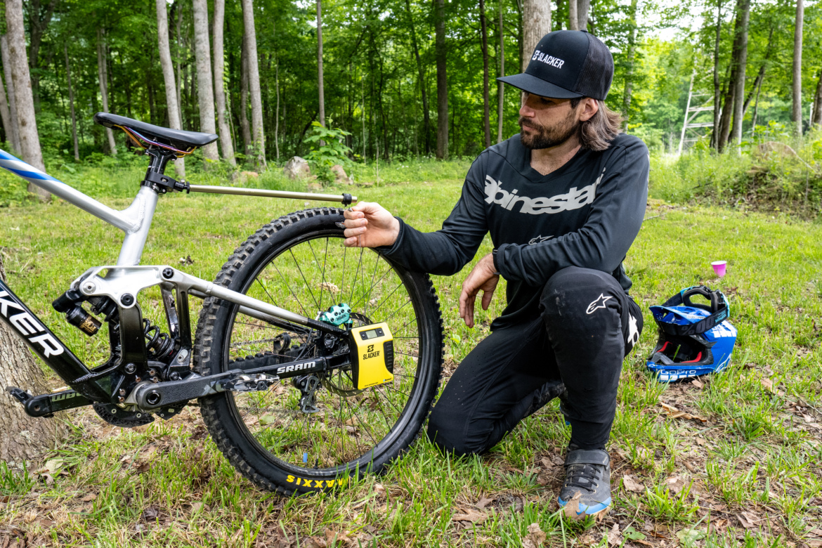 A cyclist in black gear kneels beside a mountain bike, inspecting the rear wheel and drivetrain components in a forested area. The bike features a prominent logo and a yellow device attached to it. A blue helmet rests on the ground nearby.