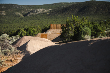 A landscape view featuring sandy mounds and vegetation, with wooden structures partially visible in the background against a backdrop of rolling green hills. The image captures a natural setting bathed in sunlight. Picuris Pueblo Bike Park mountain bike trail.