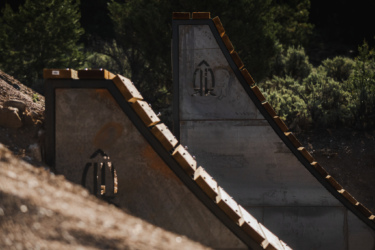 Concrete ramps designed for skateboarding or biking, featuring wooden edges, set against a backdrop of greenery and earth. The image shows two ramps positioned side by side, with a focus on their smooth surfaces and geometric shapes. Picuris Pueblo Bike Park mountain bike trail.