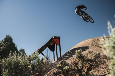 A cyclist performing a mid-air jump off a dirt ramp, surrounded by desert vegetation and a clear blue sky. Picuris Pueblo Bike Park mountain bike trail.