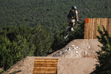 A mountain biker in mid-air performing a jump over a dirt ramp, with a backdrop of dense greenery and rocky terrain. The scene captures the excitement of biking in an outdoor park. Picuris Pueblo Bike Park mountain bike trail.