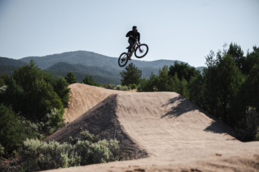 A mountain biker performing a jump over a dirt ramp in a natural outdoor setting, with mountains and trees in the background under a clear blue sky. Picuris Pueblo Bike Park mountain bike trail.