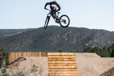 A mountain biker performing a jump off a wooden ramp, with a backdrop of mountains and clear blue sky. The rider is seen mid-air, showcasing an impressive aerial maneuver. Surrounding vegetation includes shrubs in the foreground. Picuris Pueblo Bike Park mountain bike trail.