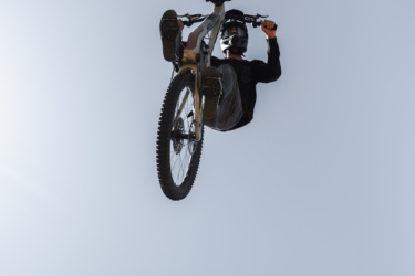 A cyclist performing a mid-air stunt on a mountain bike, suspended upside down above a wooden ramp against a clear blue sky. Picuris Pueblo Bike Park mountain bike trail.