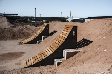 Two wooden ramps are positioned on a dirt surface, with one ramp on the left and another on the right. The ramps are constructed with slatted wooden surfaces and are supported by concrete bases. In the background, there are mounds of dirt and some industrial structures, with a clear blue sky above. Picuris Pueblo Bike Park mountain bike trail.