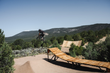 A cyclist performing a mid-air jump above a wooden ramp in a mountainous area, surrounded by trees and under a clear blue sky. Picuris Pueblo Bike Park mountain bike trail.