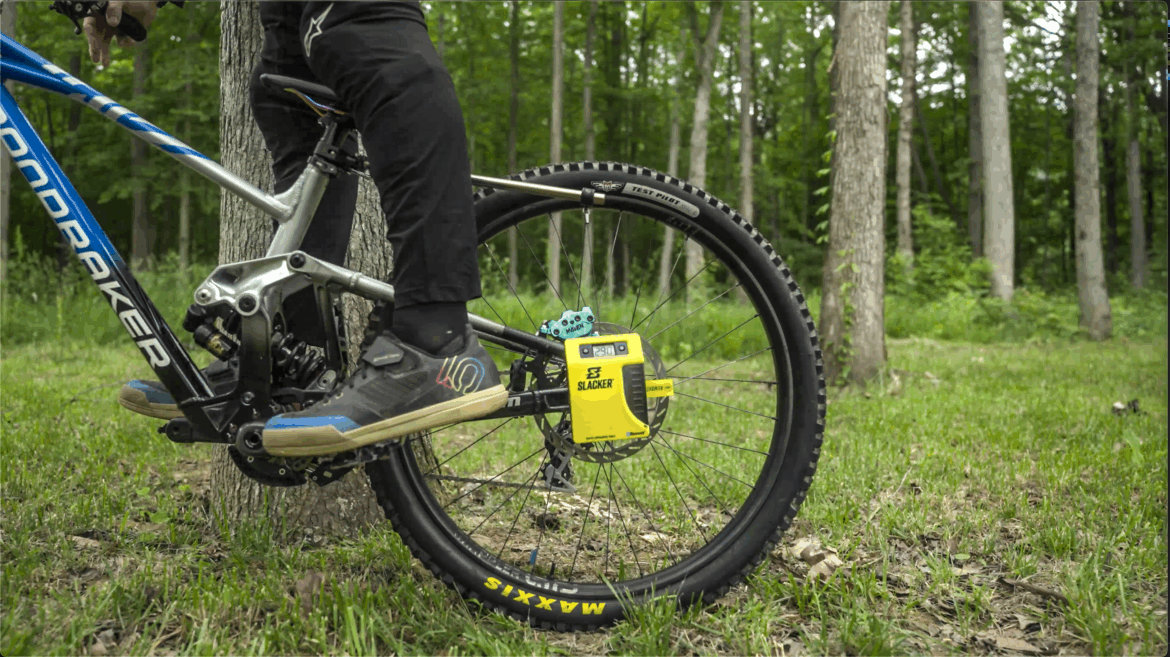 A close-up view of a mountain bike's rear wheel and the rider's foot on the pedal, set in a forested area with green grass and trees in the background. The bike features a blue frame with visible branding, and there is a yellow gadget attached to the wheel's hub. The rider is wearing dark pants and specialized biking shoes.