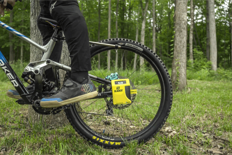A close-up view of a mountain bike's rear wheel and the rider's foot on the pedal, set in a forested area with green grass and trees in the background. The bike features a blue frame with visible branding, and there is a yellow gadget attached to the wheel's hub. The rider is wearing dark pants and specialized biking shoes.