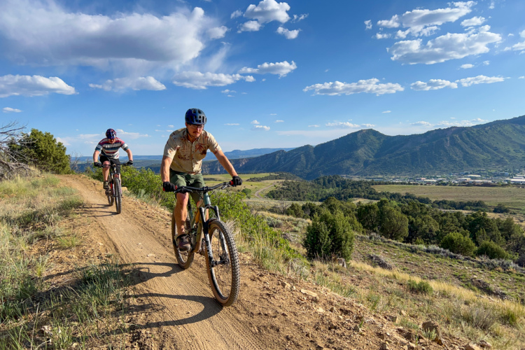 Two mountain bikers ride along a dirt trail in a scenic outdoor landscape. The background features rolling hills and a clear blue sky with scattered clouds. One cyclist is in the foreground, wearing a helmet and a light-colored shirt, while the other rides slightly behind, sporting a striped jersey. Lush greenery and rocky terrain frame the trail, enhancing the natural beauty of the setting.