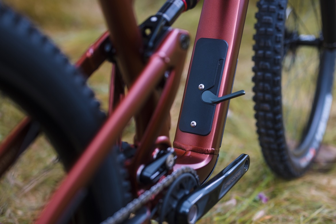 Close-up view of a mountain bike frame showcasing a metallic red color, with a focus on the bottom bracket area and a black protective cover. The image highlights details like the frame's construction and pedal crank assembly, set against a blurred natural background.