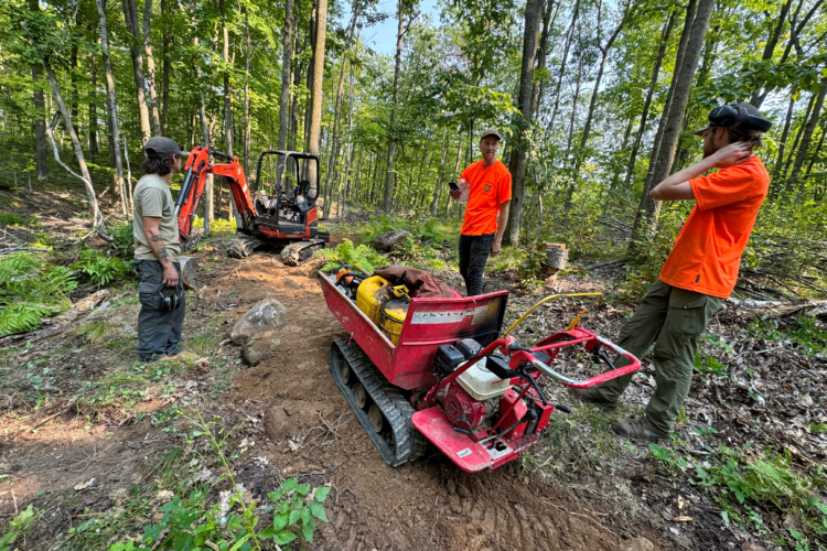 Three workers are in a forested area, engaged in trail building or maintenance. One worker stands with a tool, while two others, wearing bright orange shirts, converse near a motorized cart filled with equipment. An excavator is visible in the background, surrounded by trees and undergrowth. The scene captures the effort involved in outdoor labor in a natural setting.