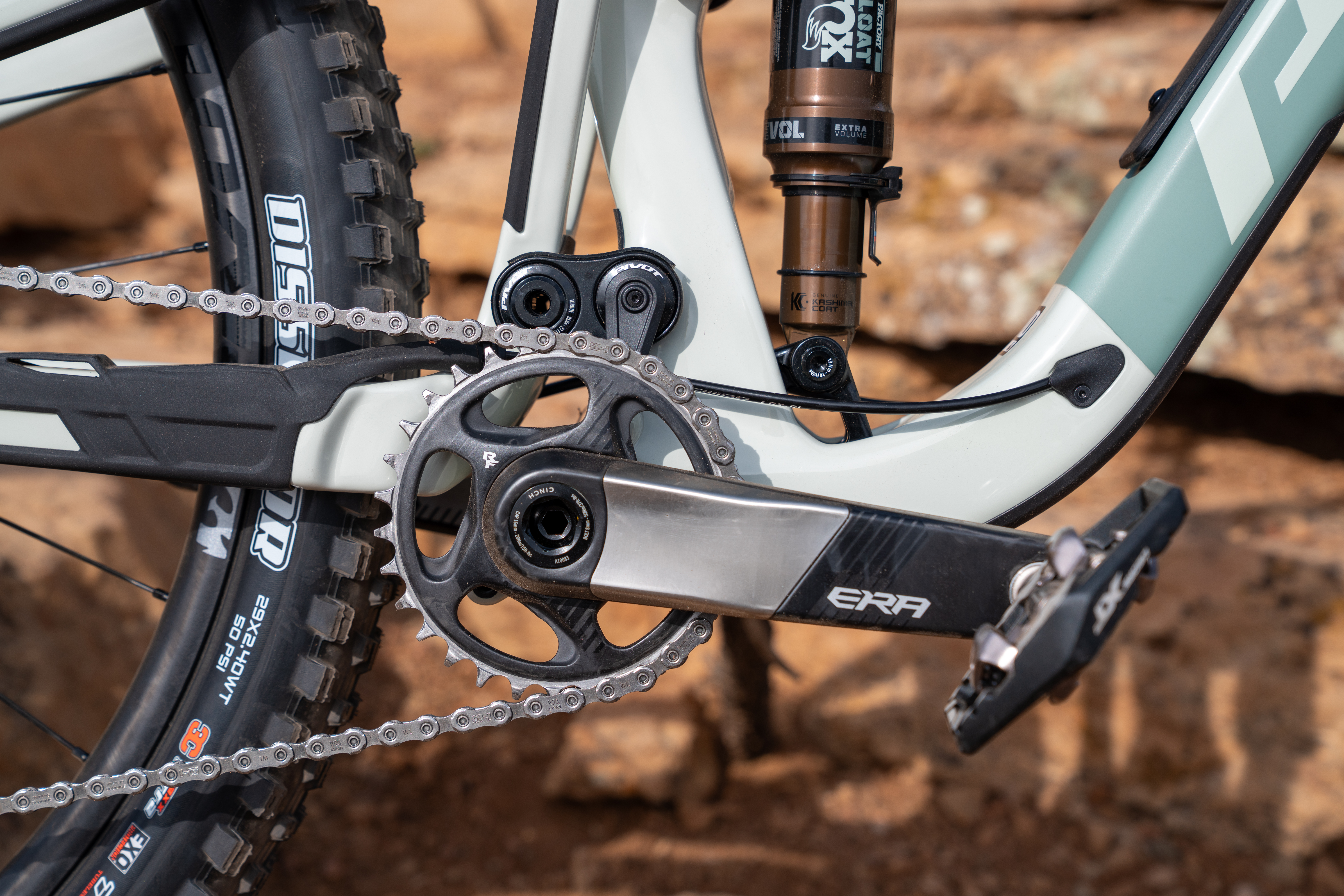 Close-up view of a mountain bike's drivetrain, featuring a chain, crankset, and pedals. The frame is a light color, and there's a visible suspension component. The background shows rocky terrain, suggesting an outdoor setting.