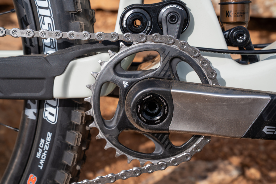 Close-up image of a mountain bike's crankset, featuring a black chainring, a silver crank arm, and a chain. The bike's tire and part of the frame are visible in the background, highlighting the rugged terrain.