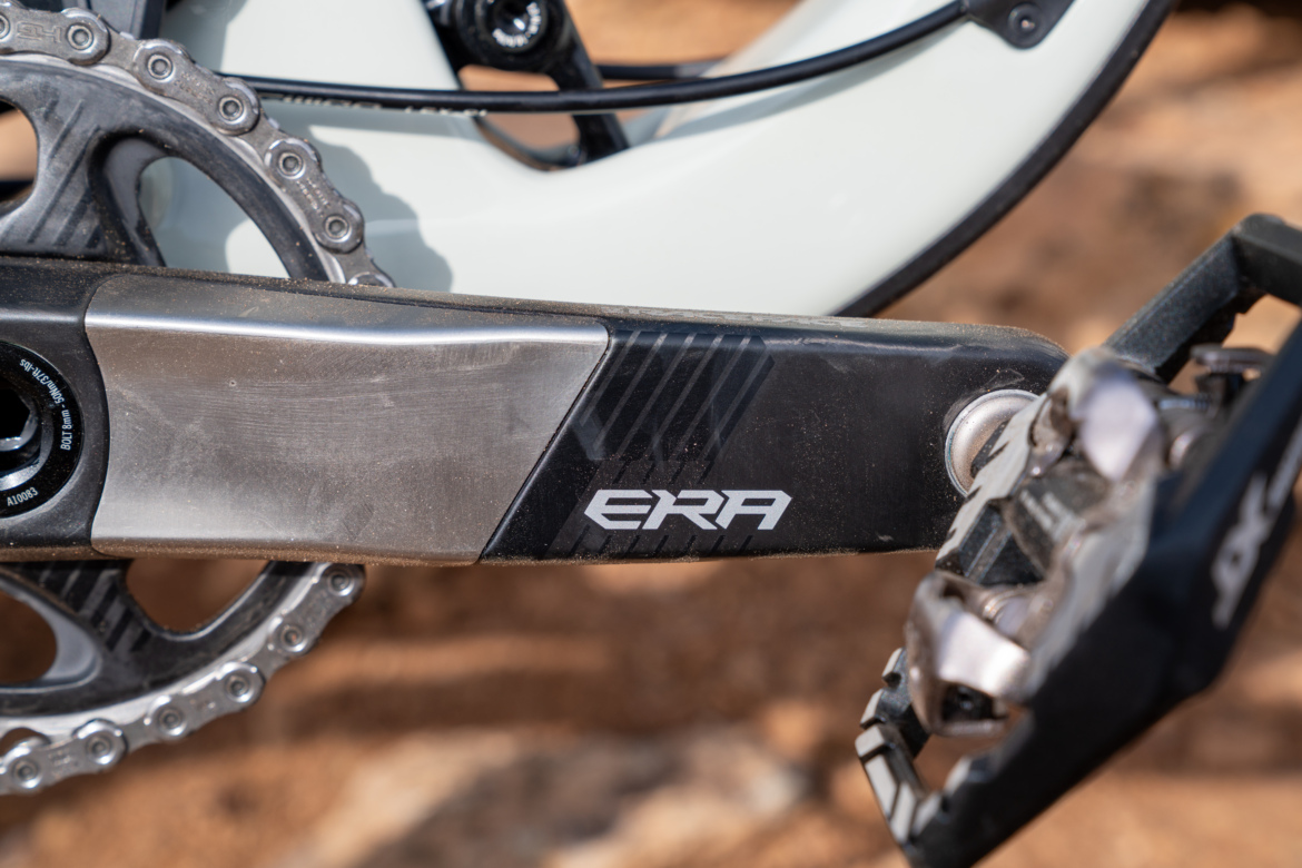 Close-up image of a bicycle crankset featuring the brand name "ERA," with a metallic finish and a visible pedal attached. The background shows a blurred, earthy terrain, suggesting an outdoor biking environment.