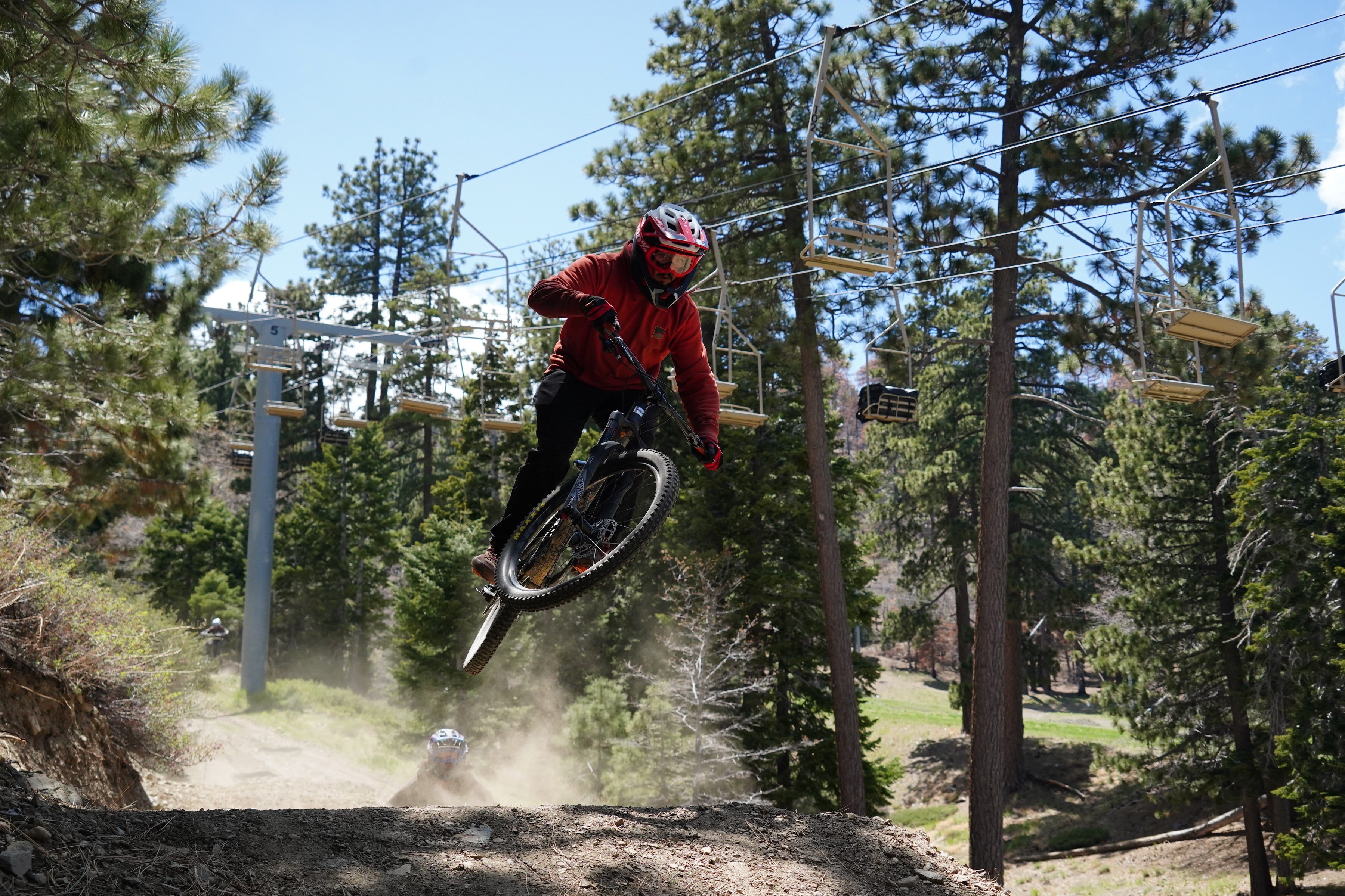 A mountain biker in a red jacket performs a jump on a dirt trail while skiing chairs are visible in the background among tall pine trees. Dust is kicked up from the bike