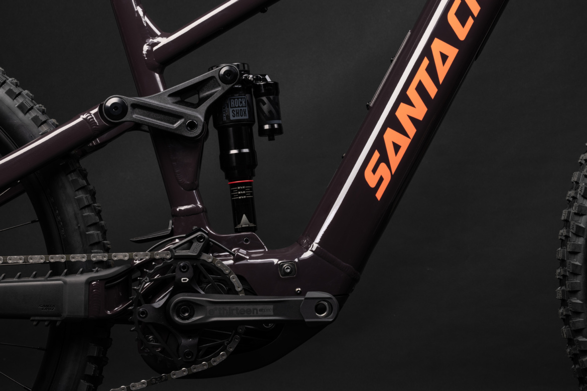 Alt tag: Close-up view of a mountain bike frame featuring the brand name "Santa Cruz," highlighting the rear suspension and drivetrain components. The frame is a deep purple color with contrasting orange logo details, set against a dark background.