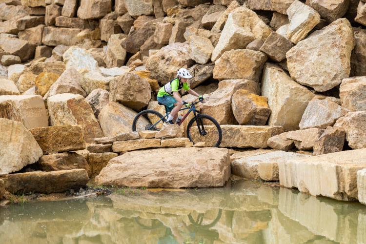 A young cyclist navigating a rocky terrain on a mountain bike, with large boulders and a small pond reflecting in the background. The cyclist is wearing a bright green shirt and a helmet, showcasing a moment of skillful riding over natural obstacles.