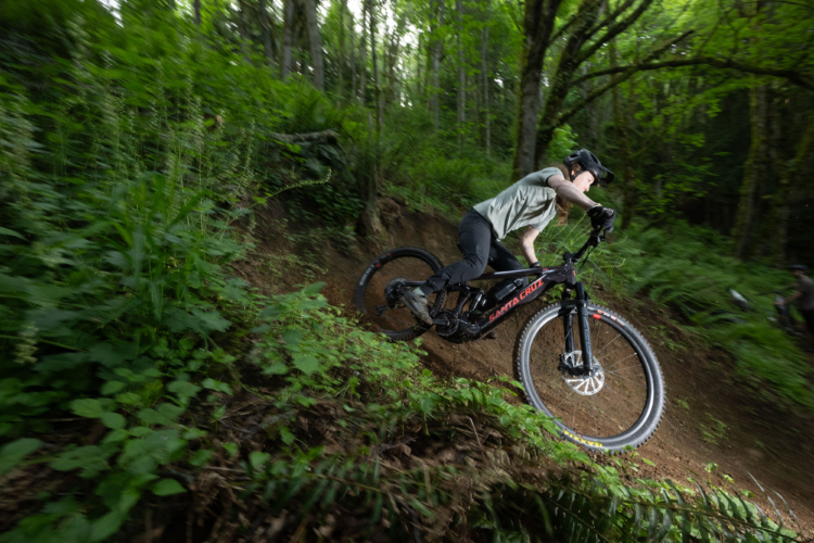 A mountain biker leans into a turn on a forested trail, surrounded by lush green foliage. The bike is partially off the ground, showcasing a dynamic action shot that captures the thrill of outdoor riding.