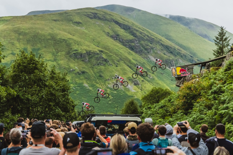 A mountain biker performing a jump off a ramp, captured in multiple stages, with a crowd of spectators watching in the foreground against a backdrop of green hills and cloudy skies. A Red Bull sign is visible near the ramp.