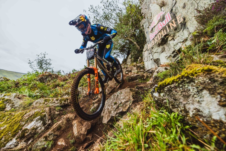 A mountain biker in a blue and black riding outfit navigates a rocky trail, surrounded by lush greenery and boulders. The rider is focused and leaning forward, with a helmet and goggles for safety. A Red Bull logo is visible on a rock in the background, indicating the location of a biking event. The sky is overcast, suggesting a cool, adventurous atmosphere.