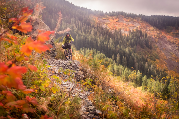 A cyclist navigating a rocky trail in a mountainous area, surrounded by vibrant autumn foliage. The scene features a backdrop of lush green trees and orange and red leaves, set against a cloudy sky.