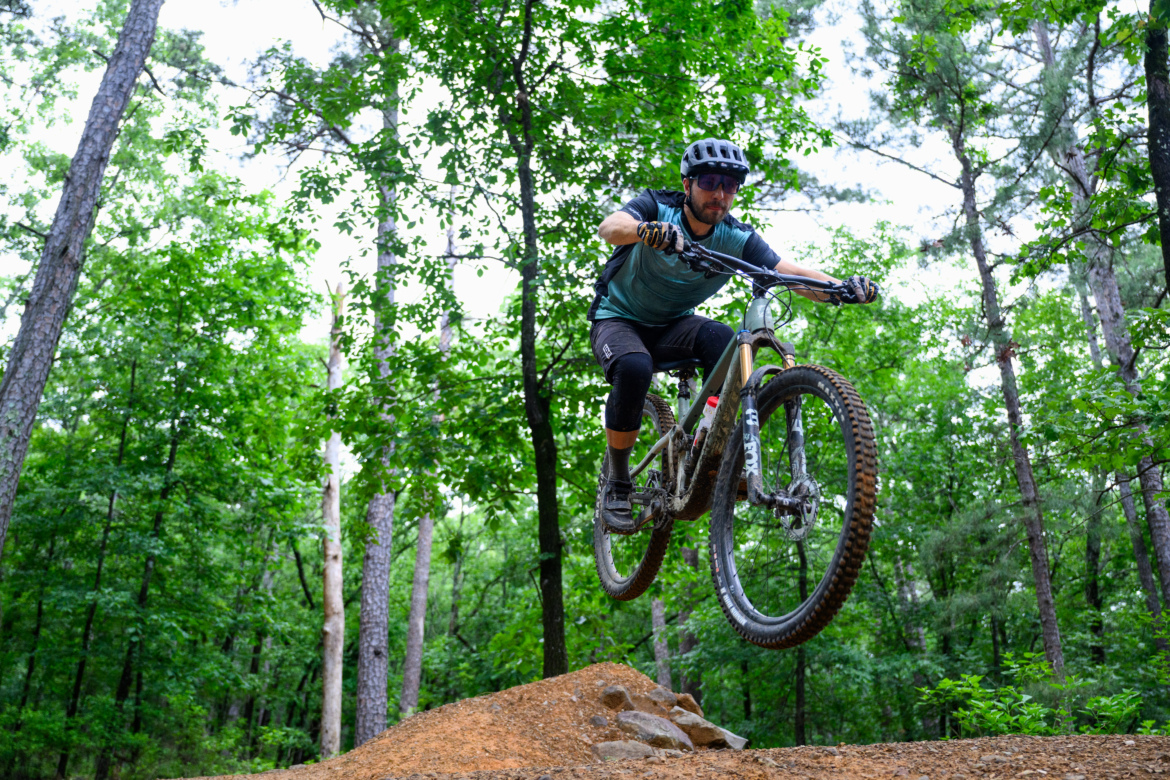 A mountain biker mid-air, performing a jump over a dirt ramp in a lush, green forest. The cyclist is wearing a helmet and protective gear, showcasing an adventurous moment in an outdoor setting.