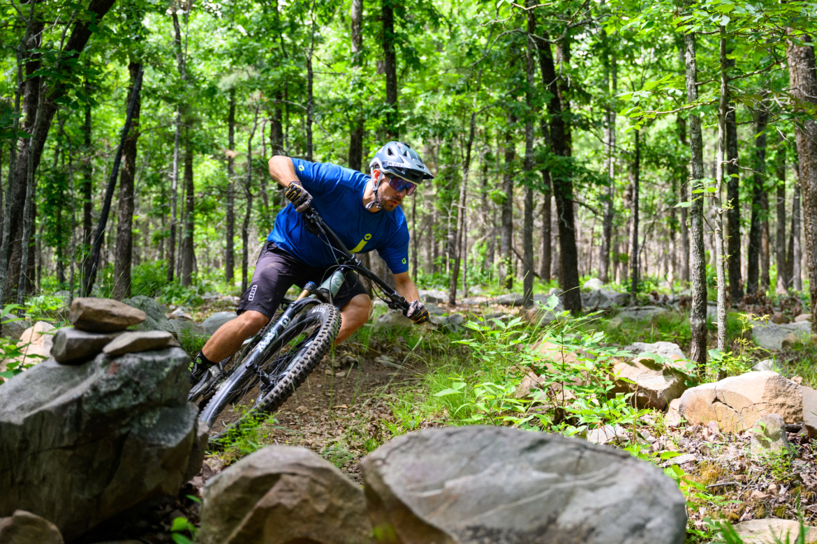 A mountain biker navigating a rocky trail through a dense forest, leaning into a turn while wearing a helmet and protective gear. The scene is filled with lush green trees and underbrush.