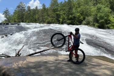 A person in biking gear is holding a mountain bike above their head while standing on a rocky surface near a waterfall, surrounded by lush green trees and a bright blue sky. The waterfall cascades in the background, creating a dynamic scene of outdoor adventure. Bridal Veil Falls mountain bike trail.
