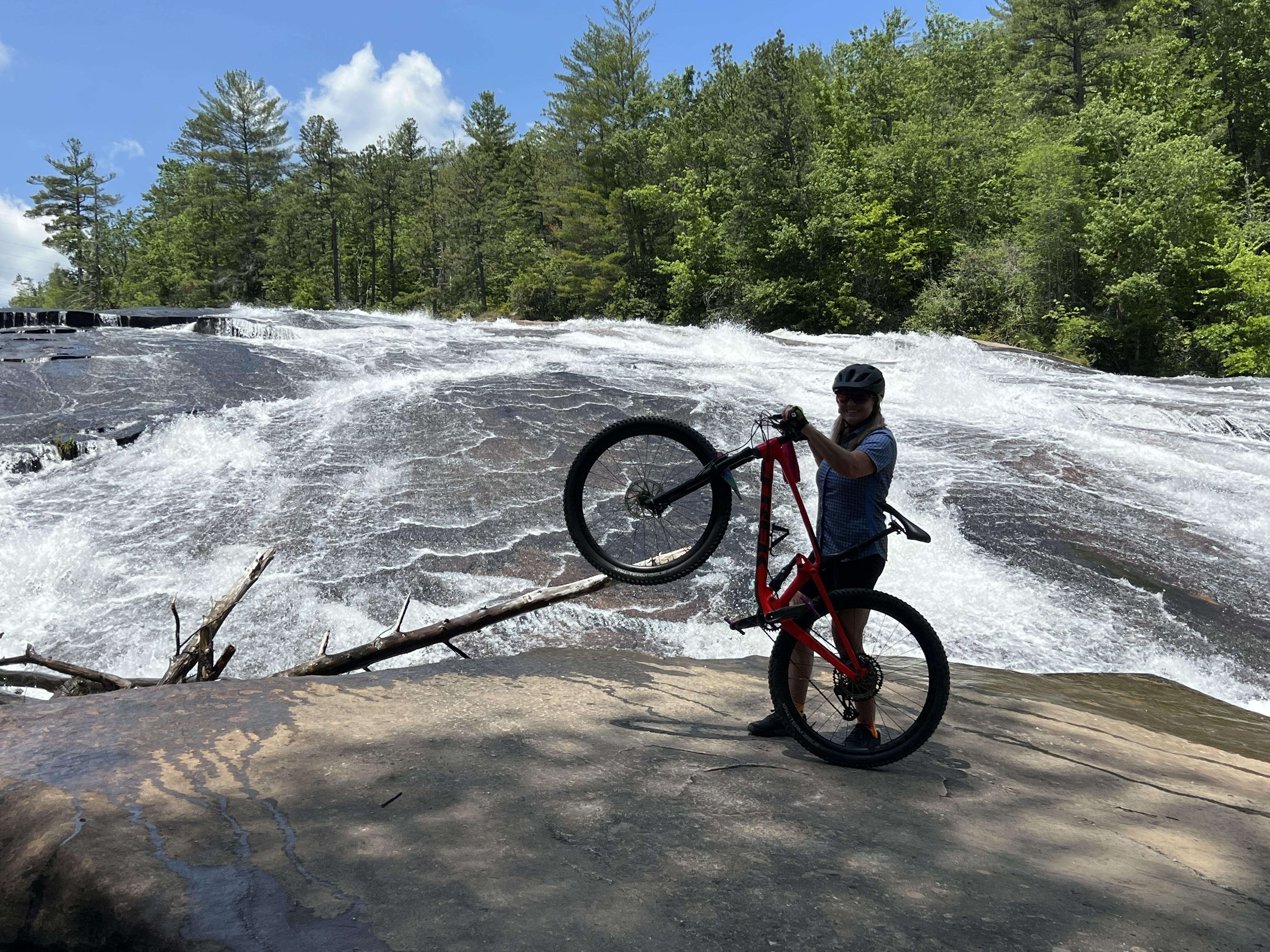 A person in biking gear is holding a mountain bike above their head while standing on a rocky surface near a waterfall, surrounded by lush green trees and a bright blue sky. The waterfall cascades in the background, creating a dynamic scene of outdoor adventure. Bridal Veil Falls mountain bike trail.