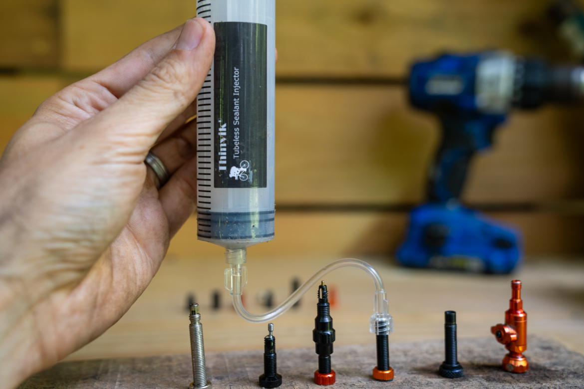 A hand holding a tubeless sealant injector next to various valve stems and a cordless drill in a workshop setting.