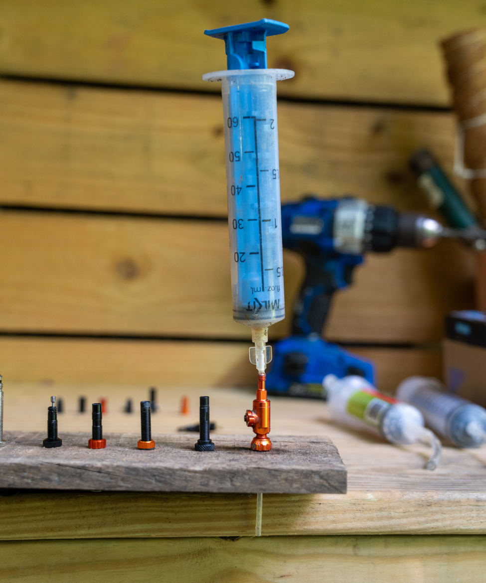 A blue syringe filled with liquid is positioned above an orange component, with several black and orange fasteners arranged on a wooden surface. In the background, a blue power drill is visible along with other syringes. The setting appears to be a workspace or workshop.