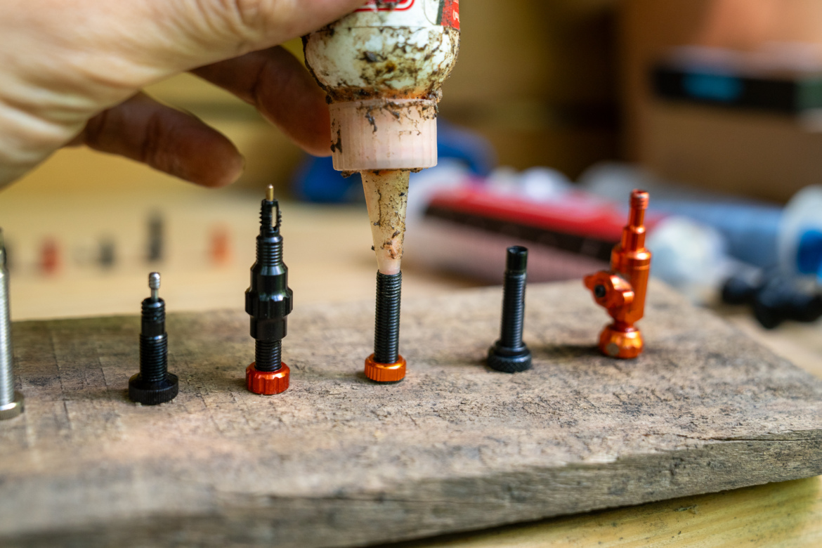 A hand holding a bottle of lubricant, dispensing it onto a threaded metal bolt, with several bolts and fittings arranged on a wooden surface in the background.