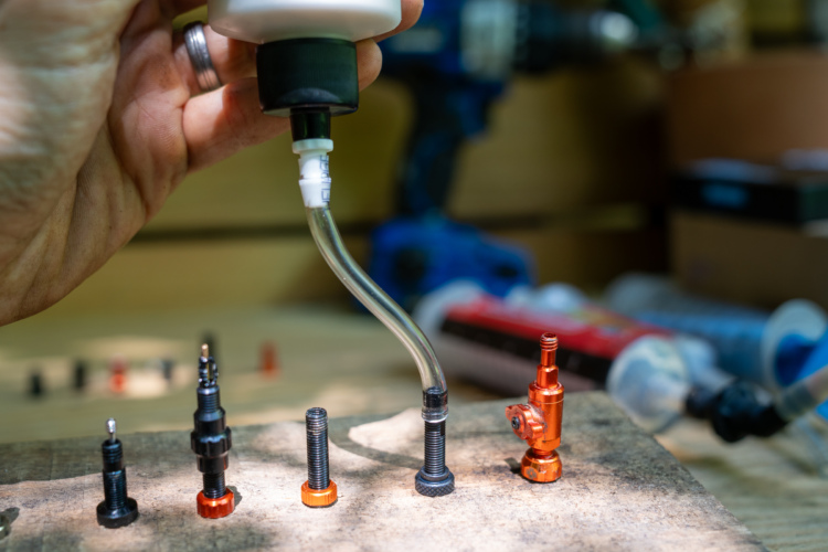 A hand holding a squeeze bottle with a clear tube extends toward a variety of small metal parts and screws arranged on a wooden surface. In the background, tools and containers are partially visible, suggesting a workshop setting. The focus is on the precision components, including some with orange accents.