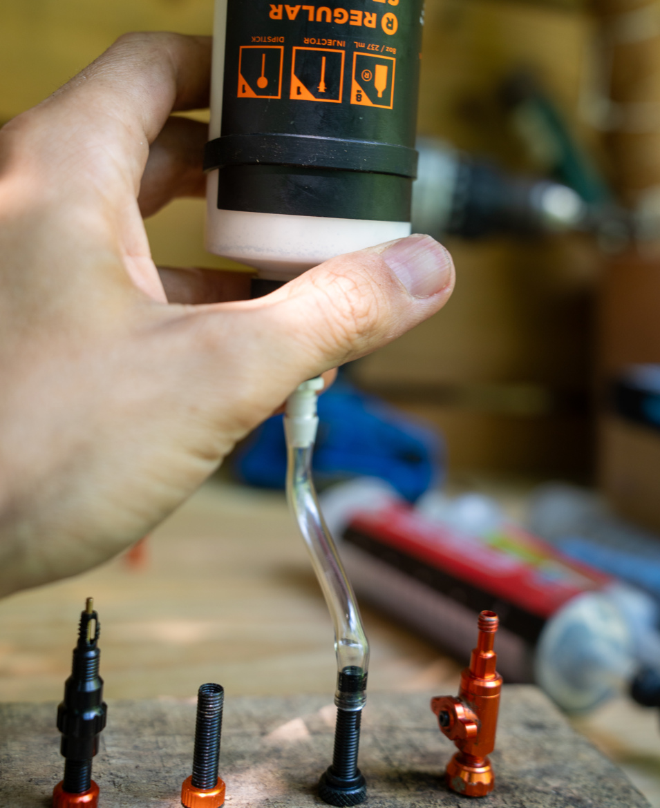 A hand holds a bottle of lubricant with a clear tube attached, positioned above a series of metallic parts, including several black and orange components, on a wooden work surface. The background is slightly blurred, with tools and other objects out of focus.