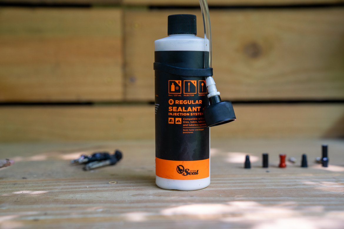 A bottle of sealant for tire repair, with a clear plastic tube attached to the top and a nozzle cap. The bottle is labeled "Regular Sealant Injection System" and is sitting on a wooden surface, alongside various small nozzle adapters.