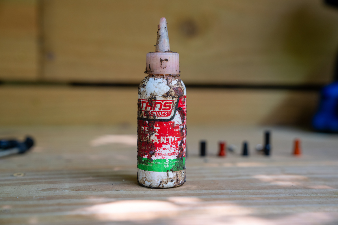 A weathered bottle of tire sealant with a pink nozzle, sitting on a wooden surface. The label is partially faded, and the bottle has dirt and grime on it. In the background, there are several small valve caps in various colors, including black, red, and orange.