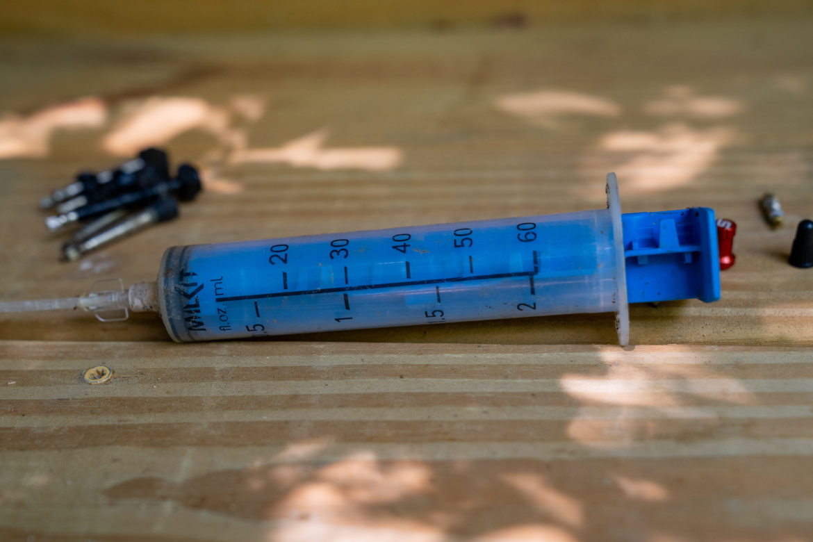 A plastic syringe with measurement markings and a blue plunger resting on a wooden surface, accompanied by several small black and metallic objects in the background.