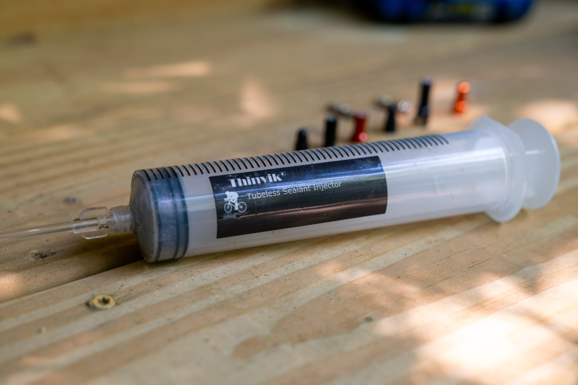 A tubeless sealant injector resting on a wooden surface, with multiple small valve cores of various colors in the background. The injector features measurement markings and a label indicating it is a "Tubeless Sealant Injector."