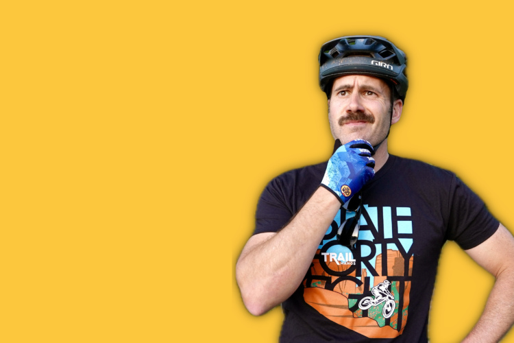 A man wearing a black t-shirt with a colorful graphic design and a mountain biking helmet is thoughtfully posing with his hand on his chin. He has a mustache and is wearing blue gloves. The background is a vibrant yellow.